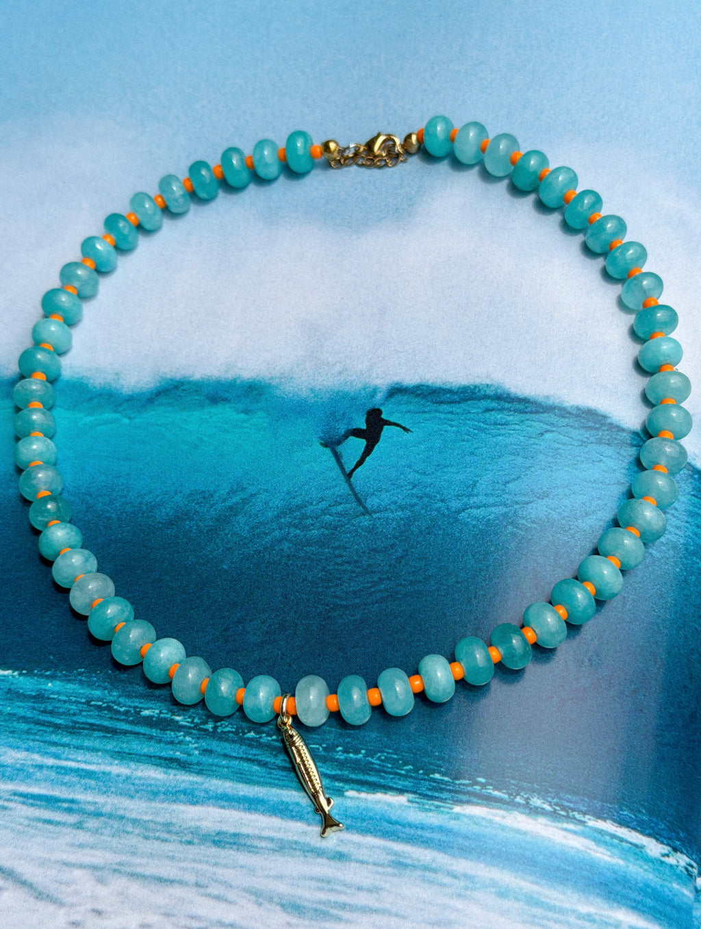 Necklace with beads and fish charm over a painting of a surfer in the ocean.