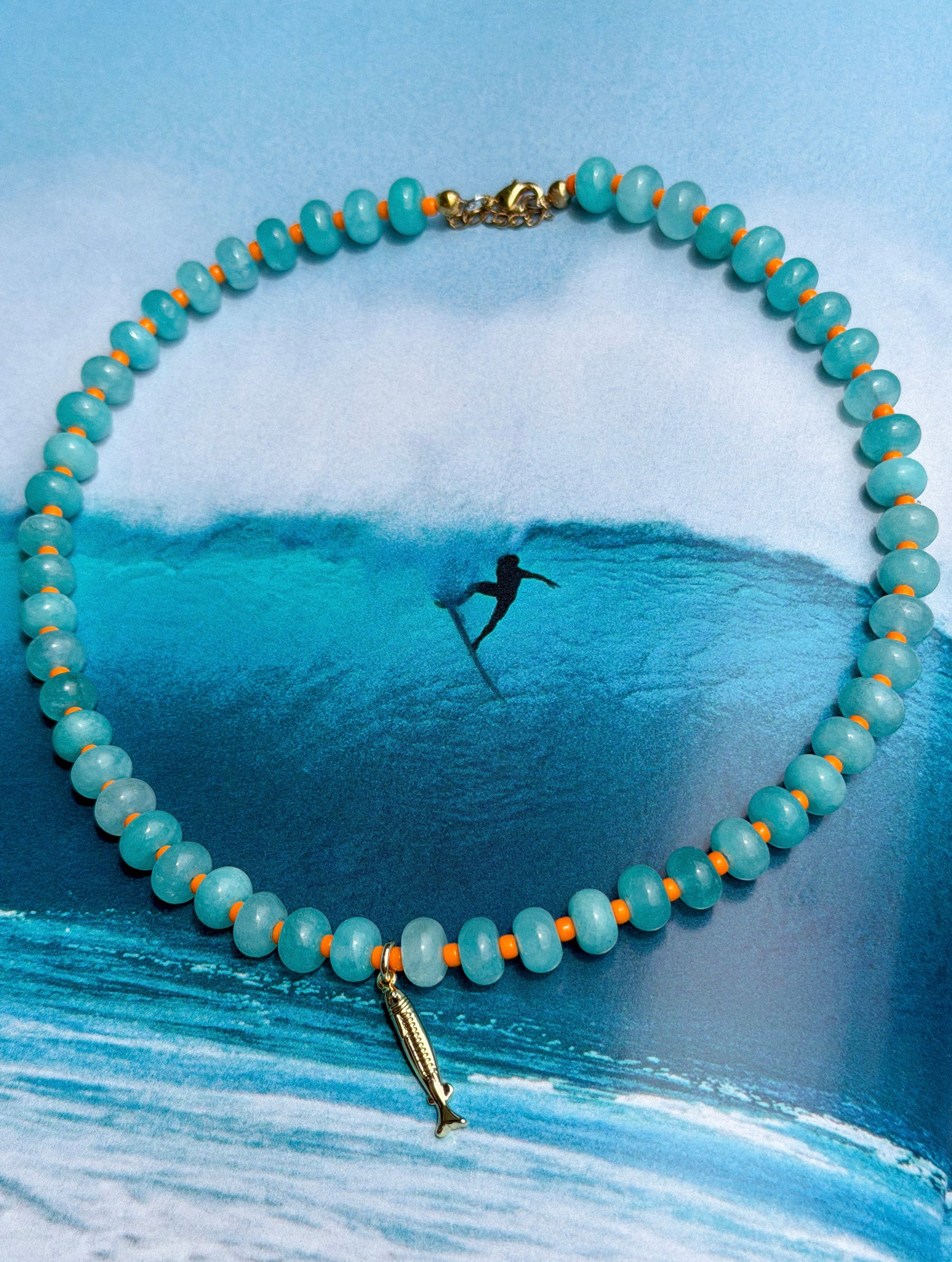 Necklace with beads and fish charm over a painting of a surfer in the ocean.